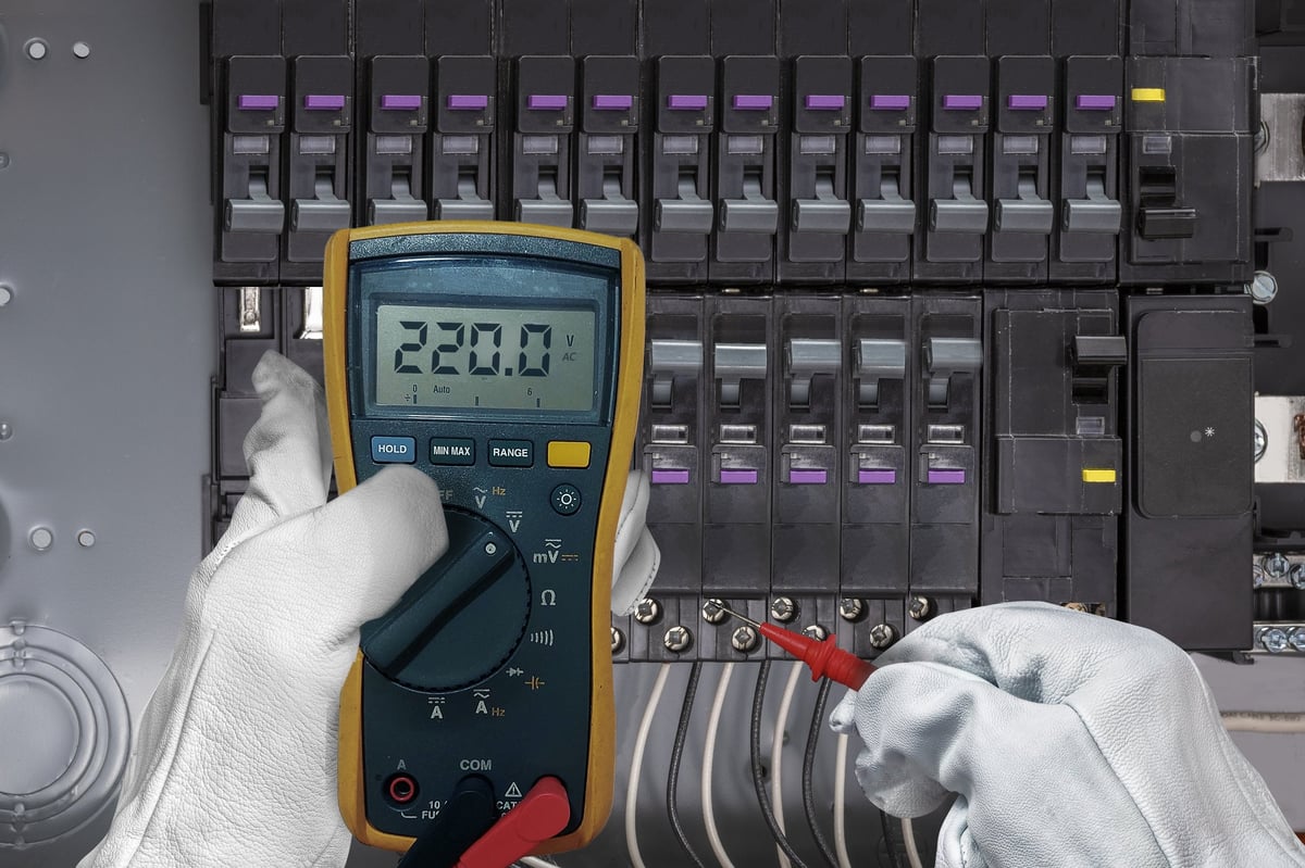Close-up of an electrician wearing protective gloves measuring 220V AC voltage using a digital multimeter inside an electrical circuit breaker panel