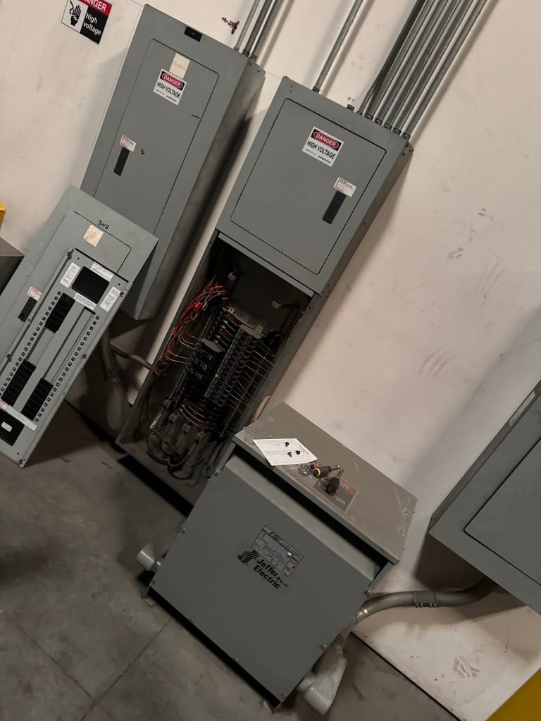 Electrical panels and switchgear mounted on white wall with exposed wiring and conduit in a utility room