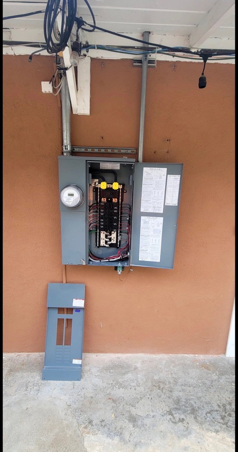 Electrical panel box mounted on orange wall with circuit breakers, meter, and overhead electrical wiring in garage